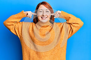 Beautiful redhead woman putting fingers on ears ignoring noise winking looking at the camera with sexy expression, cheerful and