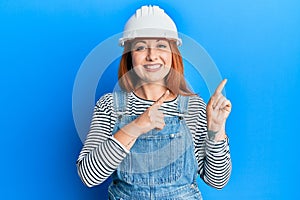 Beautiful redhead woman wearing architect hardhat smiling and looking at the camera pointing with two hands and fingers to the