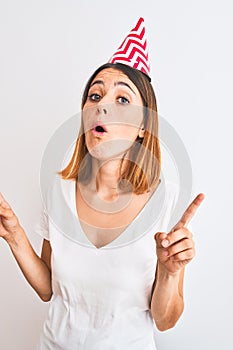 Beautiful redhead woman wearing birthday cap over isolated background amazed and surprised looking up and pointing with fingers