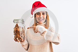 Beautiful redhead woman wearing christmas hat holding cookies jar over isolated background very happy pointing with hand and