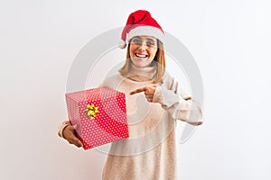 Beautiful redhead woman wearing christmas hat holding present over isolated background very happy pointing with hand and finger