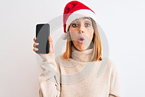 Beautiful redhead woman wearing christmas hat showing smartphone screen scared in shock with a surprise face, afraid and excited