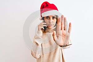 Beautiful redhead woman wearing christmas hat talking on smartphone with open hand doing stop sign with serious and confident