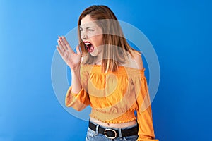 Beautiful redhead woman wearing orange casual t-shirt standing over  blue background shouting and screaming loud to side