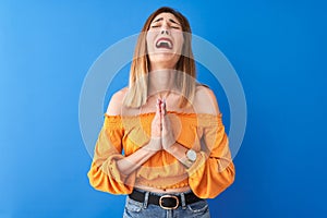 Beautiful redhead woman wearing orange casual t-shirt standing over isolated blue background begging and praying with hands