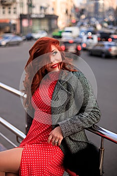 Beautiful redhead woman wearing red dress and stylish coat on city street. loneliness and depression concept