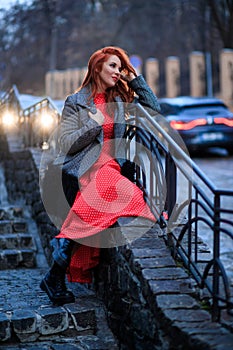 Beautiful redhead woman wearing red dress and stylish coat on city street. loneliness and depression concept