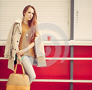 Beautiful redheaded girl with a fashionable bag in the hand is worth near the closed outdoor cafe