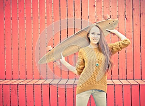 Beautiful redheaded girl holding a longboard standing near red wall