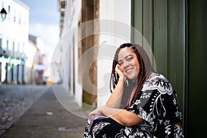 Beautiful, relaxed woman traveling with braids in her hair sitting on a step with her hand on her chin. Old houses in the