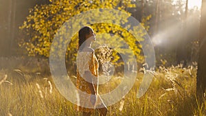 beautiful relaxed woman in yellow dress with open shoulders walking in pine forest during golden colored autumn sunset