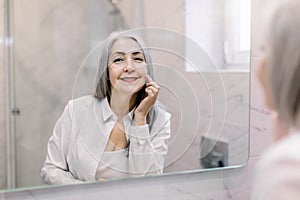 Beautiful retired gray haired lady standing in bathroom in front of the mirror, looking at camera with smile, while