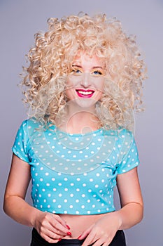 Beautiful retro-style blonde girl with voluminous curly hairstyle, in a blue polka-dot blouse on a gray background, smiles and