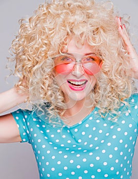 Beautiful retro-style blonde girl with voluminous curly hairstyle, in a blue polka-dot blouse and pink glasses on a gray