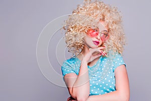 Beautiful retro-style blonde girl with voluminous curly hairstyle, in a blue polka-dot blouse and pink glasses on a gray