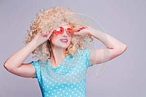 Beautiful retro-style blonde girl with voluminous curly hairstyle, in a blue polka-dot blouse and pink glasses on a gray