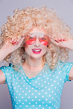 Beautiful retro-style blonde girl with voluminous curly hairstyle, in a blue polka-dot blouse and pink glasses on a gray