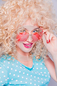 Beautiful retro-style blonde girl with voluminous curly hairstyle, in a blue polka-dot blouse and pink glasses on a gray