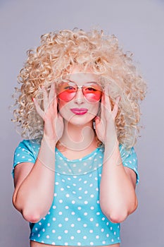 Beautiful retro-style blonde girl with voluminous curly hairstyle, in a blue polka-dot blouse and pink glasses on a gray