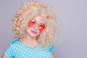 Beautiful retro-style blonde girl with voluminous curly hairstyle, in a blue polka-dot blouse and pink glasses on a gray