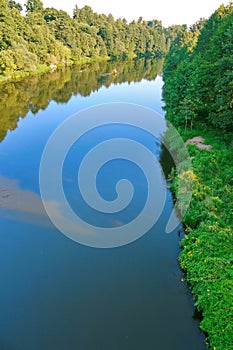 Beautiful river with a watery surface reflecting the trees growing on the shore and clouds in the sky. A good place to