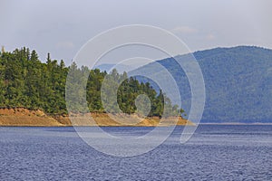 A beautiful rocky shore with tall green trees goes into the calm blue waters of the reservoir.