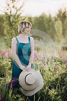 Beautiful romantic girl blonde in a dress in a field of purple flowers of lupins at dawn holds a hat in her hands