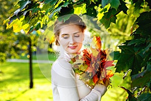 A beautiful romantic girl in a light cozy sweater with a bouquet of maple leaves in an autumn sunny park. Autumn beauty, fashion