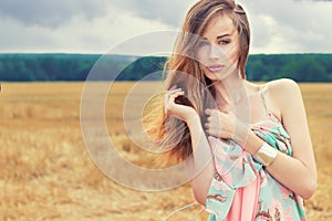 Beautiful romantic girl with red hair wearing a colored dress, the wind standing in the field on a cloudy summer day