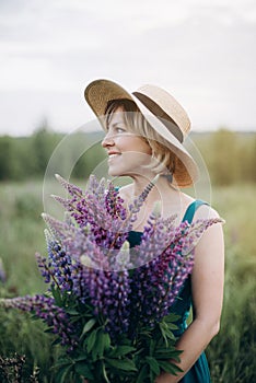 A beautiful romantic girl smiles in a dress and hat in a flower field with a huge bouquet of lupine flowers
