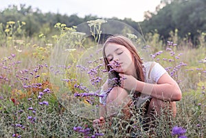 Beautiful romantic little preteen girl sitting on summer meadow enjoying aroma of wild purple oregano flowers. Summer holidays.