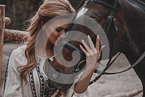 Beautiful and romantic young woman wearing the dress is embracing and stroking the horse on the ranch