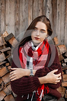 Beautiful rural young woman in stylish knitted warm clothes in a vintage scarf posing near an old wooden barn outdoors outside the
