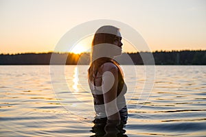 Beautiful Russian girl in a long black swimsuit swims outside the city on the lake in the rays of sunset or dawn