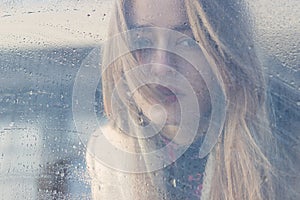 Beautiful sad girl with big eyes in a coat is behind wet glass