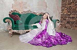 Beautiful sad woman in fantasy white and purple rococo style medieval dress sitting on the floor near sofa