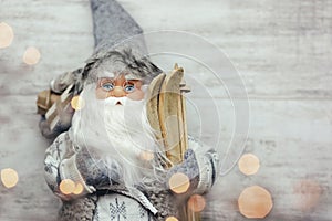 Beautiful santa claus in glasses with skis and gifts on wooden background. Blurred yellow lights on the Santa.