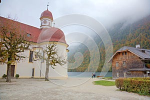 Beautiful scenery of Lake Konigssee with famous Sankt Bartholomae pilgrimage church by the lakeside and autumn mountains in foggy photo