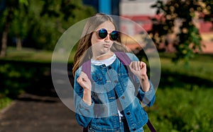 Beautiful schoolgirl girl teenager, in summer in park on nature, wearing sunglasses behind backpack and in denim jacket