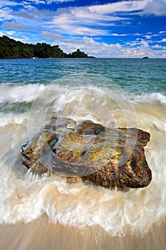 Beautiful sea coast landscape with waves and stone. Green island in the background. Big stone in the water. Ocean with dark blue