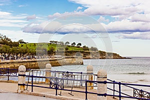 Beautiful seascape with rolling waves on a seashore behind blue metal fence on a promenade in summer day. Green hills on