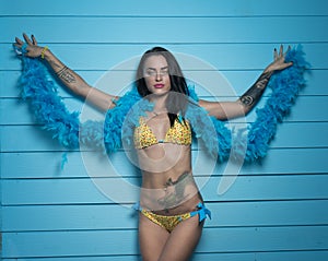 Beautiful seductive brunette woman wearing yellow bikini and blue feather boa looking into the camera and posing over blue