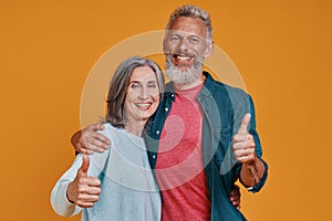 Beautiful senior couple looking at camera and keeping thumbs up while standing together against orange background