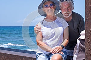 Beautiful senior couple sitting close to the sea looking at camera smiling. Two retired enjoying summer vacation and freedom