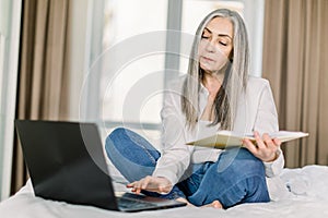 Beautiful senior gray haired woman in casual wear working on laptop computer while sitting on bed in bedroom, with her