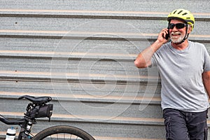 Beautiful senior man with beard talks to the phone standing against a gray wall. Resting after an healthy excursion with e bike.