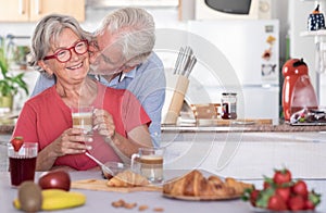 Beautiful senior man kissing his wife having breakfast at home. Retired happy people  drinking cappuccino eating fruit and