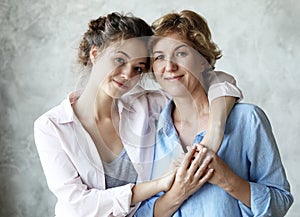 Beautiful senior mom and her adult daughter are hugging, looking at camera and smiling. At Home