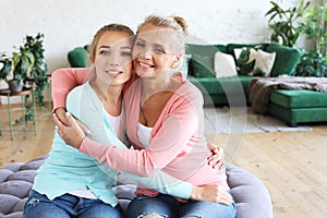Beautiful senior mom and her adult daughter are hugging, looking at camera and smiling. At Home.