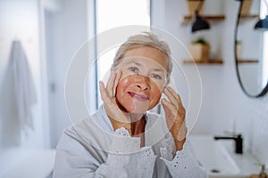 Beautiful senior woman in bathrobe, applying eye patches for puffiness while looking at camera photo